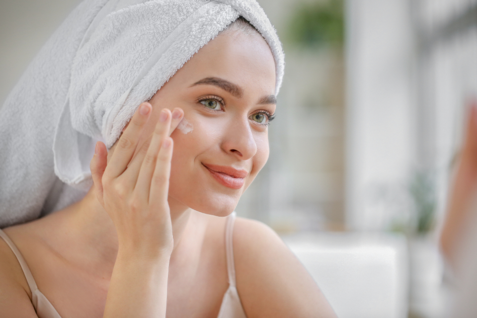 Smiling woman in a bath towel applying face cream as part of her skincare routine